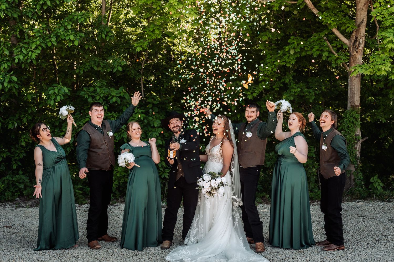 Wedding party in green dresses and brown suits celebrating outdoors, tossing confetti with lush green trees in background.