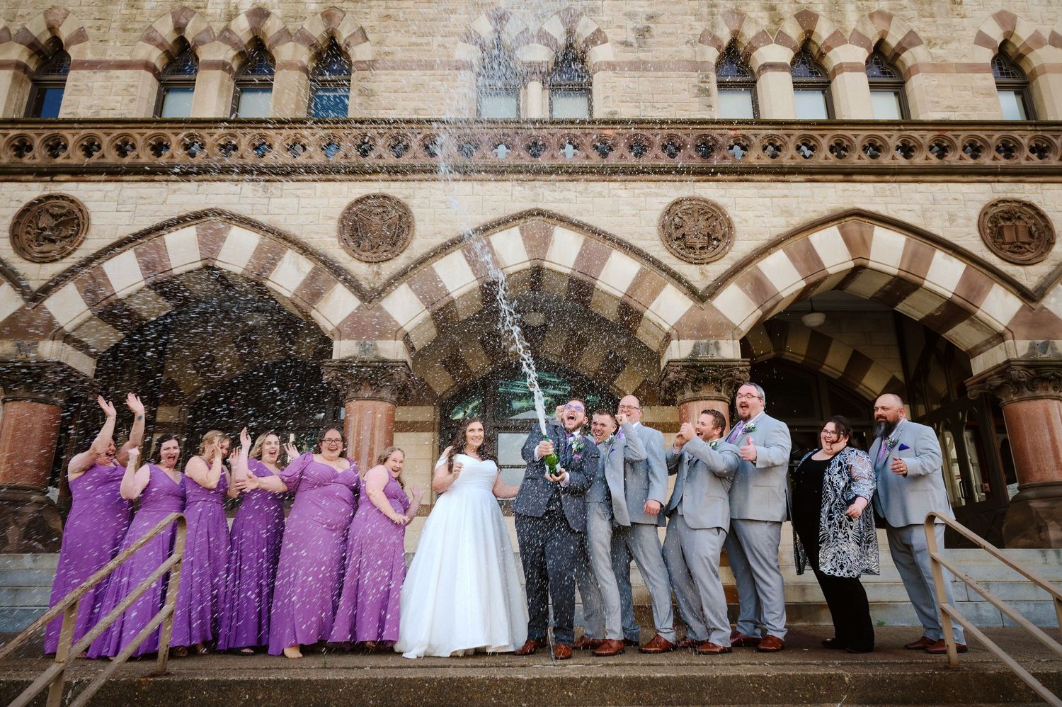 Wedding party poses in front of ornate stone architecture with bridesmaids in purple dresses and groomsmen in gray suits.