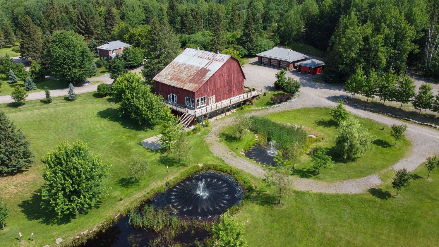 Aerial view of a red barn house with circular driveway, decorative pond and surrounding green landscape.