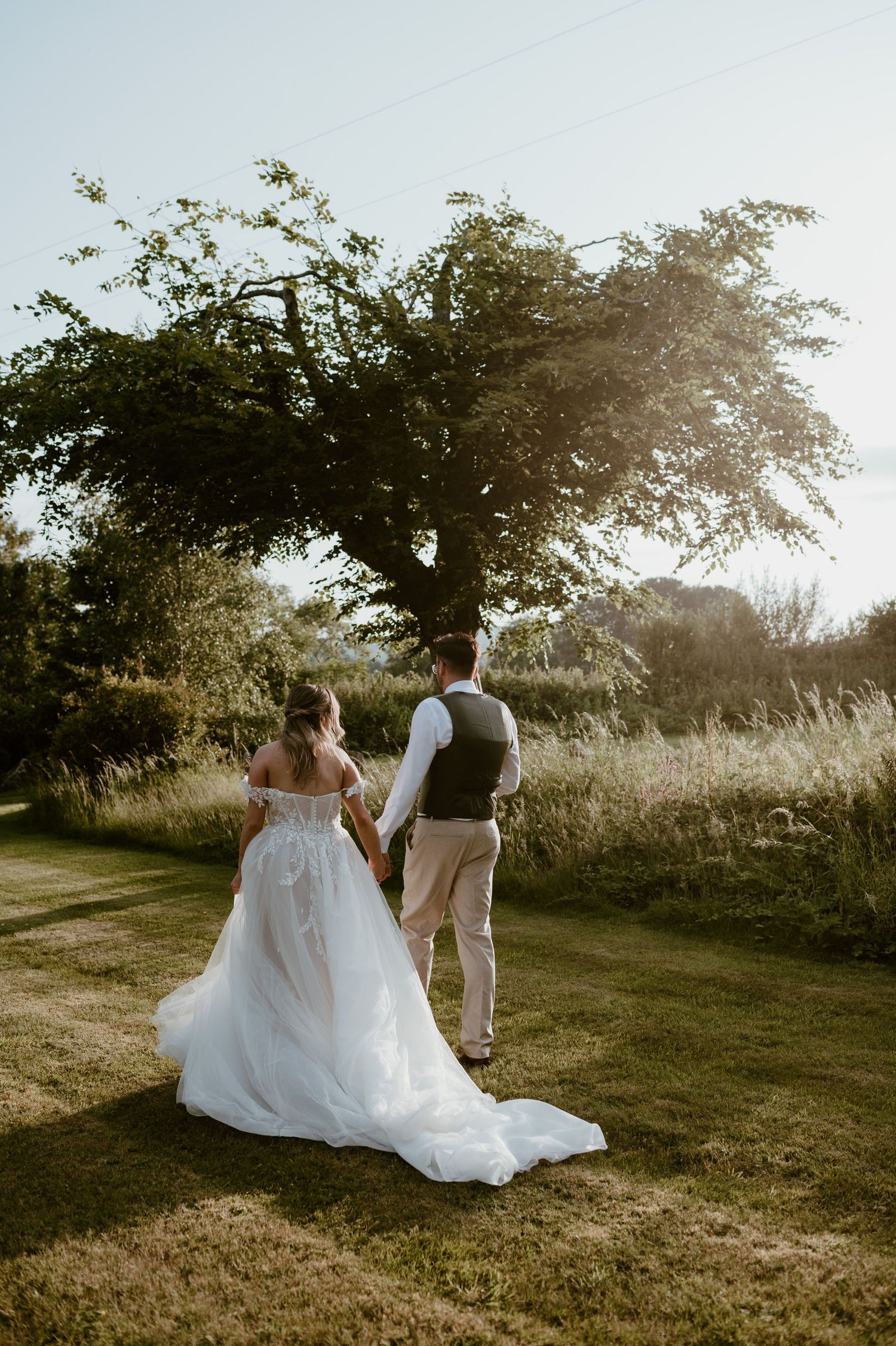A couple walks hand in hand through a sunlit meadow during golden hour as her white wedding dress trails behind them.
