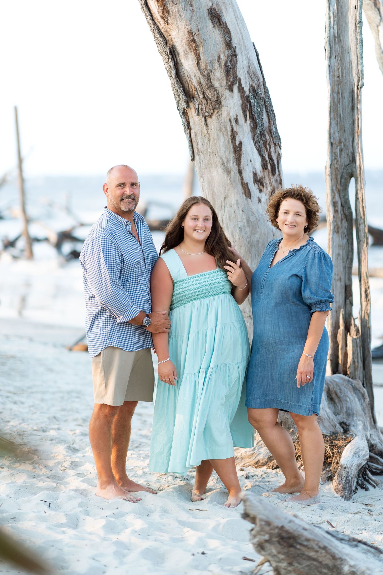 Family of three posing beside a weathered tree on during a beach family photography session in October in the South.