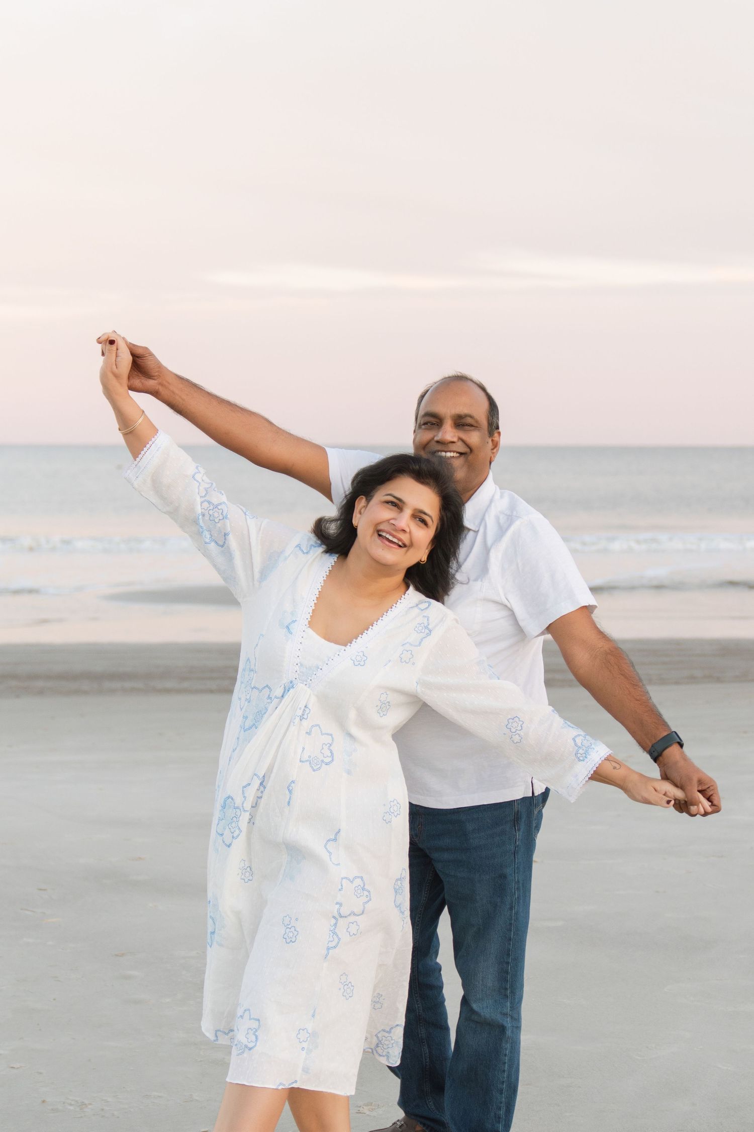 Smiling couple holding hands with arms outstretched beside the ocean during a beach portrait session at sunset in the winter.