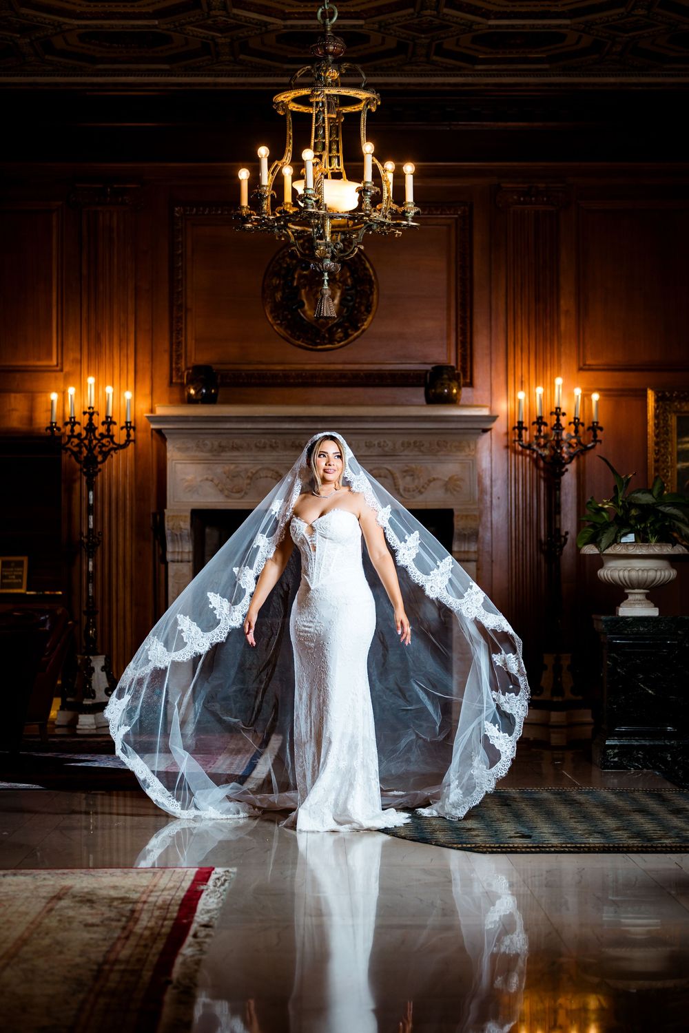 Bride in fitted white gown and lace veil poses in elegant wood-paneled room with chandelier and candelabras.