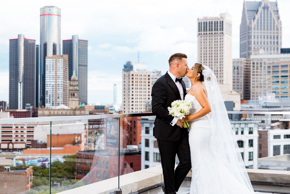 Bride and groom kissing on rooftop with Detroit city skyline backdrop, bride holding white bouquet in elegant wedding attire.
