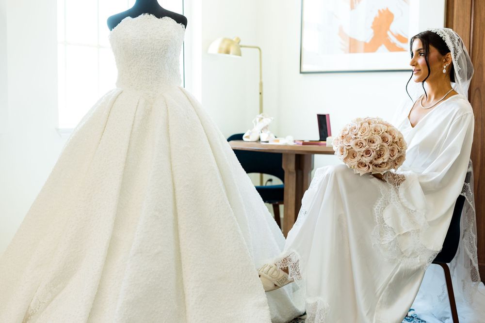 Bride in white gown holds bouquet beside elegant ballgown on mannequin in bright bridal suite.