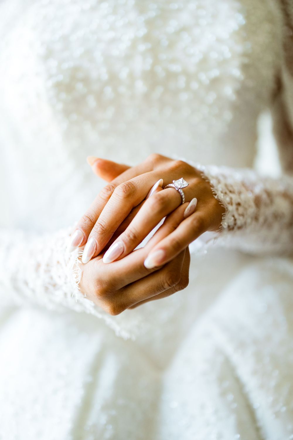Bride in white lace wedding dress clasping hands showing elegant diamond engagement ring with French manicure.