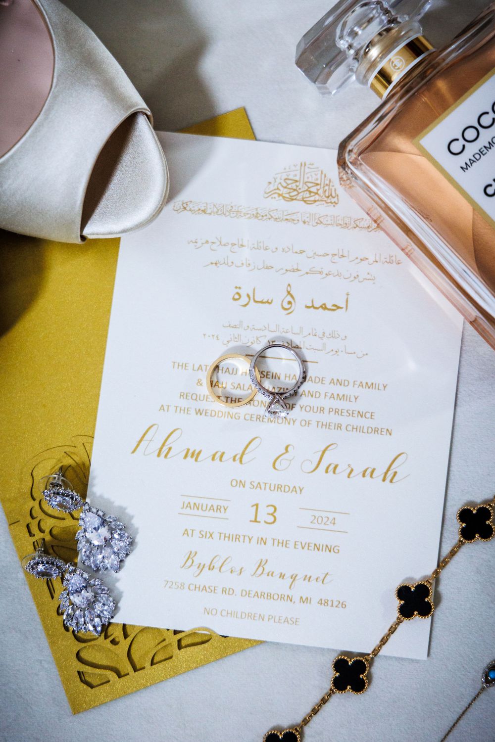 Wedding invitation in Arabic and English with gold text and wedding rings displayed on yellow paper with perfume bottle.