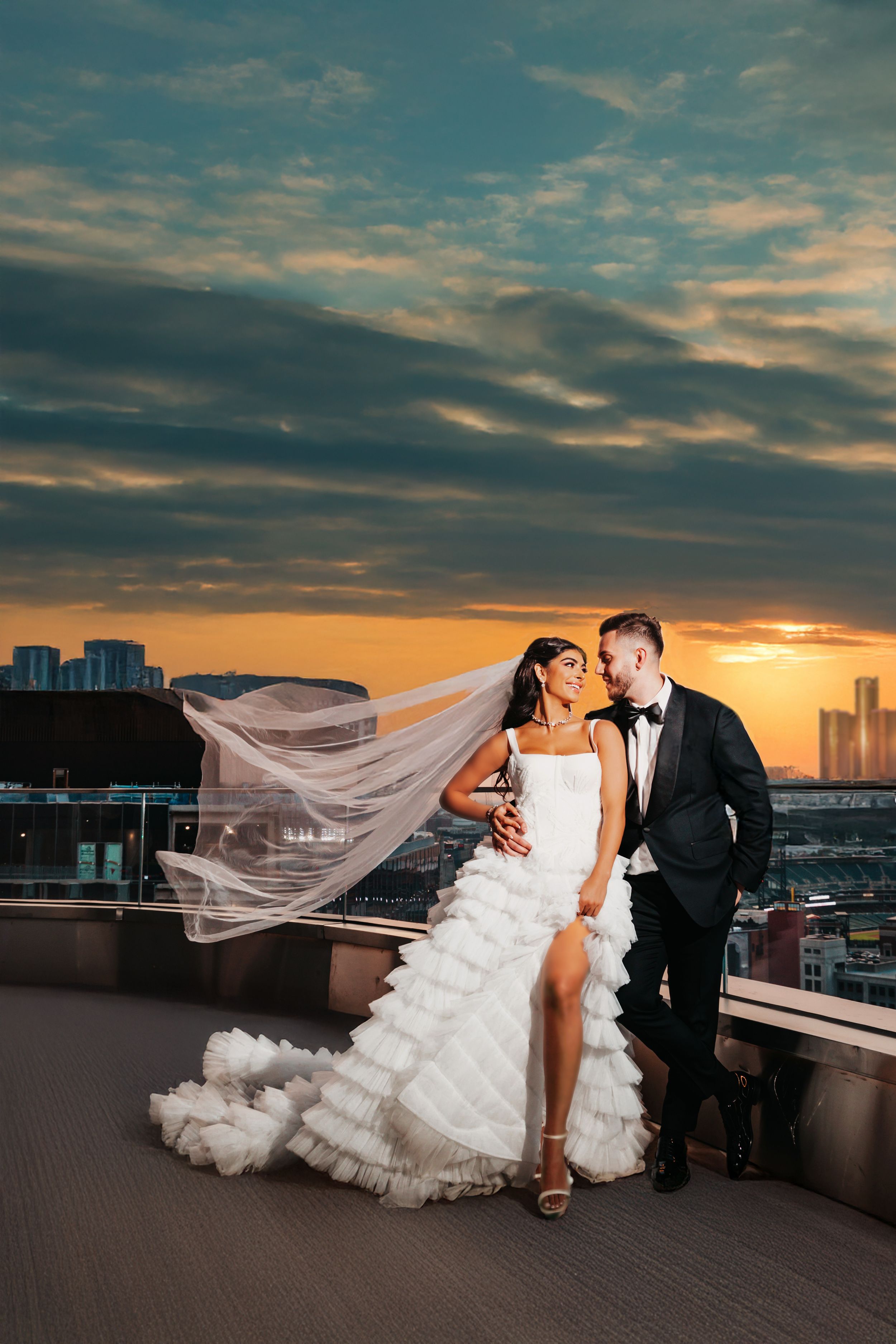 Elegant couple embraces on rooftop during sunset with flowing veil and city skyline in the background.