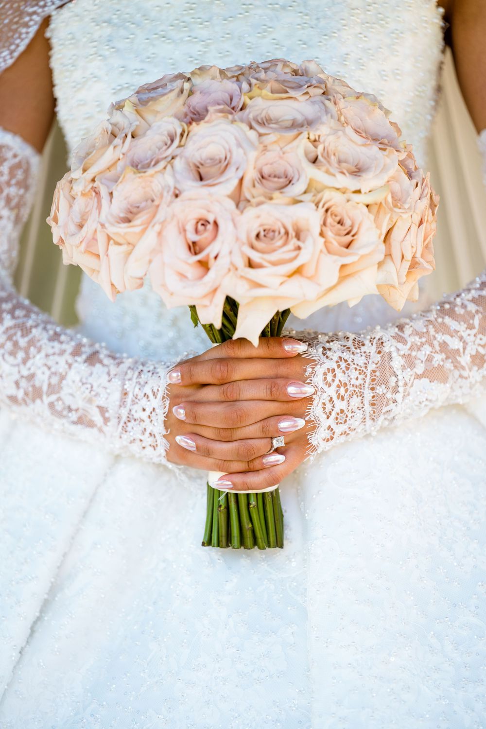 Bride in lace wedding dress holding elegant bouquet of blush pink roses with manicured hands and diamond ring.