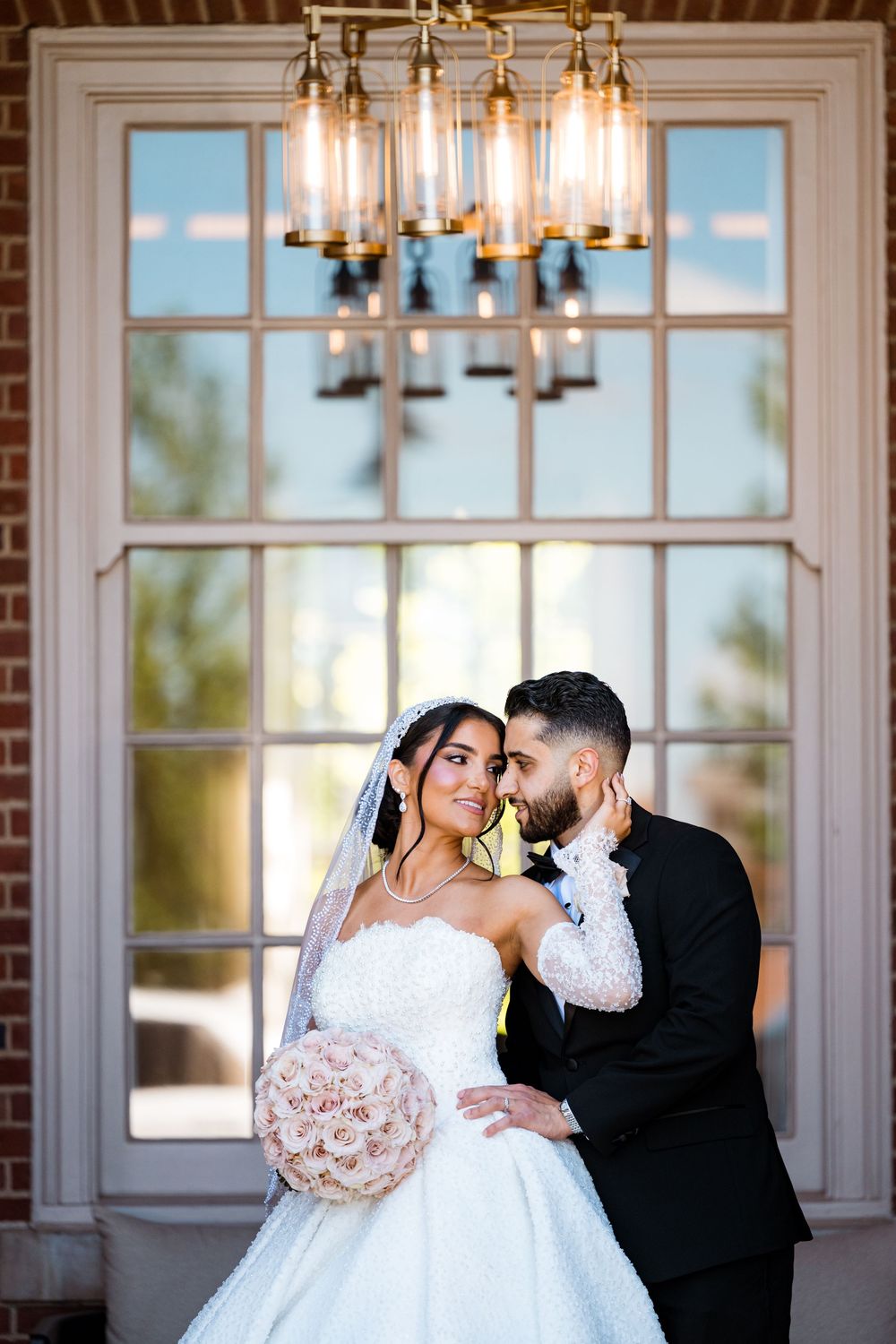 Bride in strapless lace gown holding pink bouquet with groom in tuxedo under chandelier by large window.