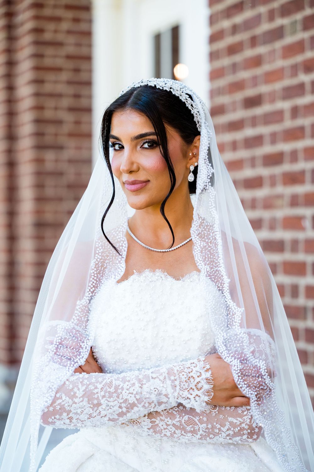 Elegant bride in white lace gown and veil with pearl tiara poses gracefully against a brick wall backdrop.