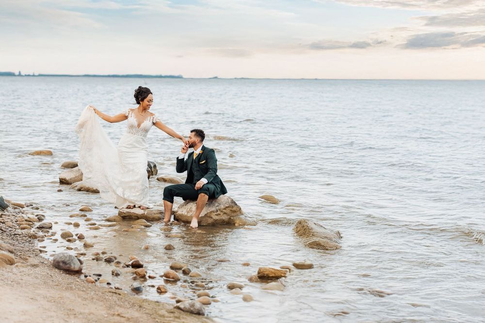 A couple in wedding attire sits on coastal rocks at the beach during sunset with waves lapping at the shore.