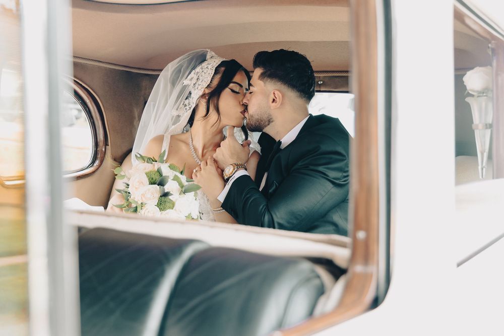 Bride and groom sharing a romantic kiss inside a vintage car on their wedding day.