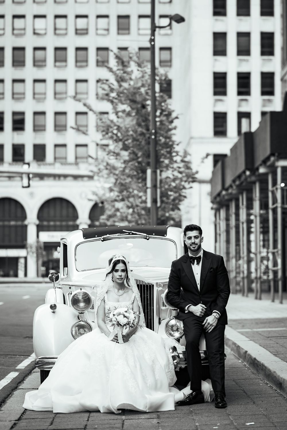 Black and white photo of bride and groom posing beside a vintage classic car on a city street.