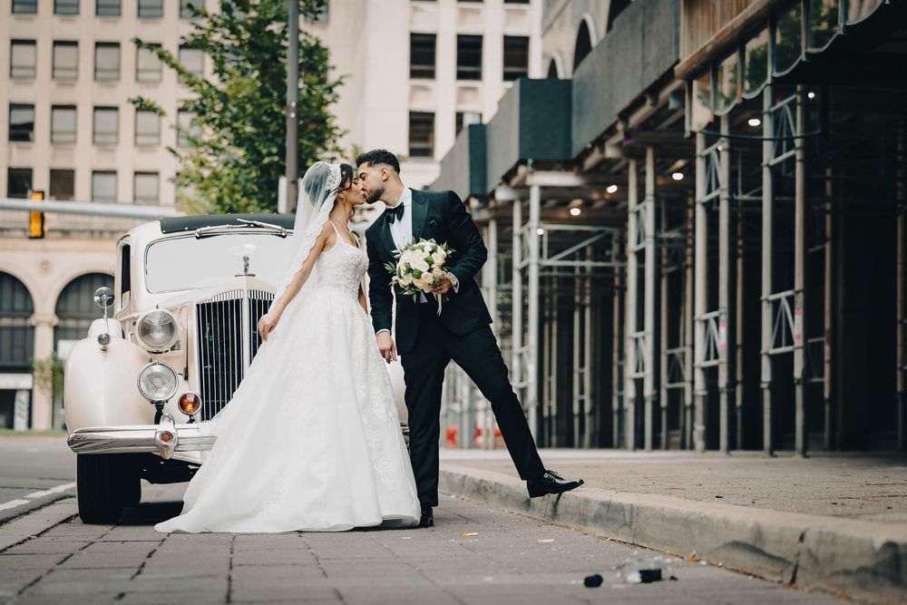 Bride and groom sharing a romantic kiss beside a vintage white car on a city street on their wedding day.