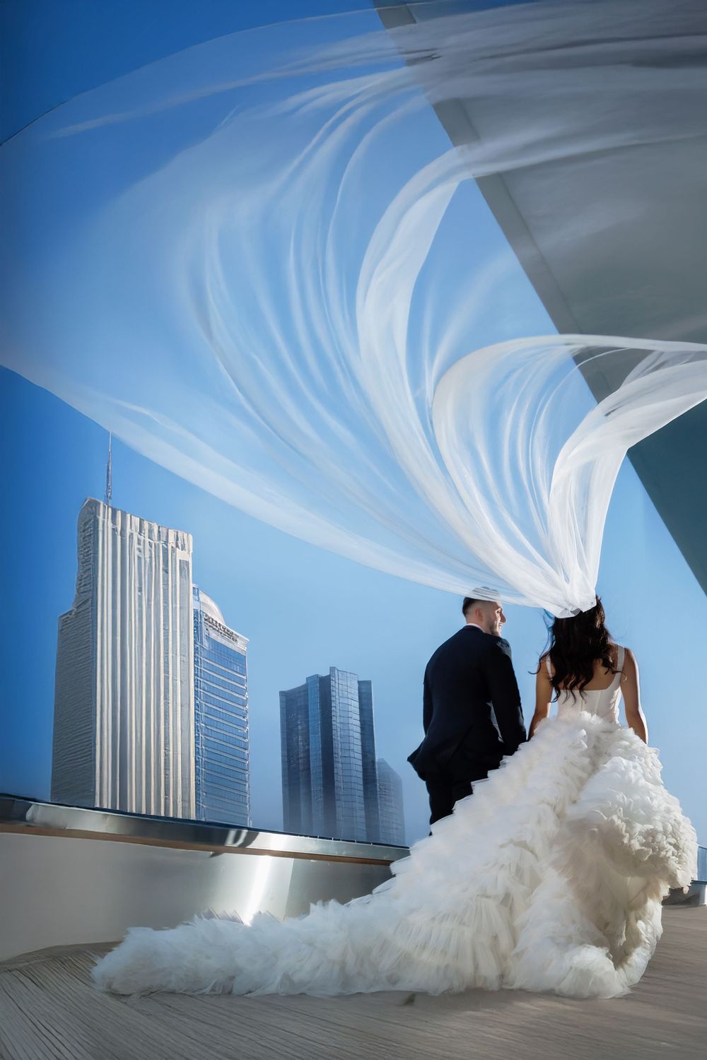 A dramatic wedding photo with a flowing veil against a modern cityscape of tall buildings under blue skies.