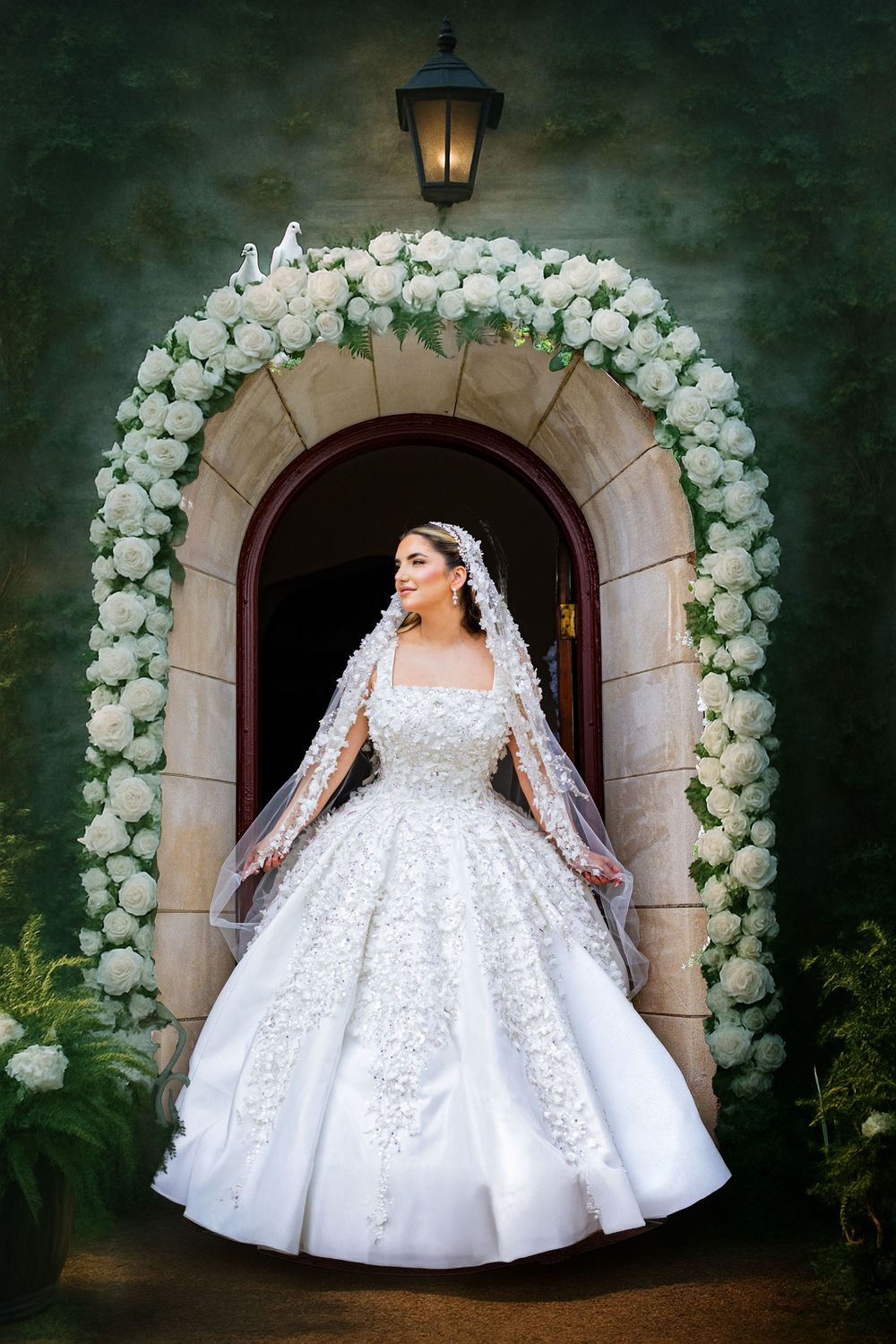 Elegant white wedding gown with lace details and veil under an archway adorned with white floral arrangements and lantern.