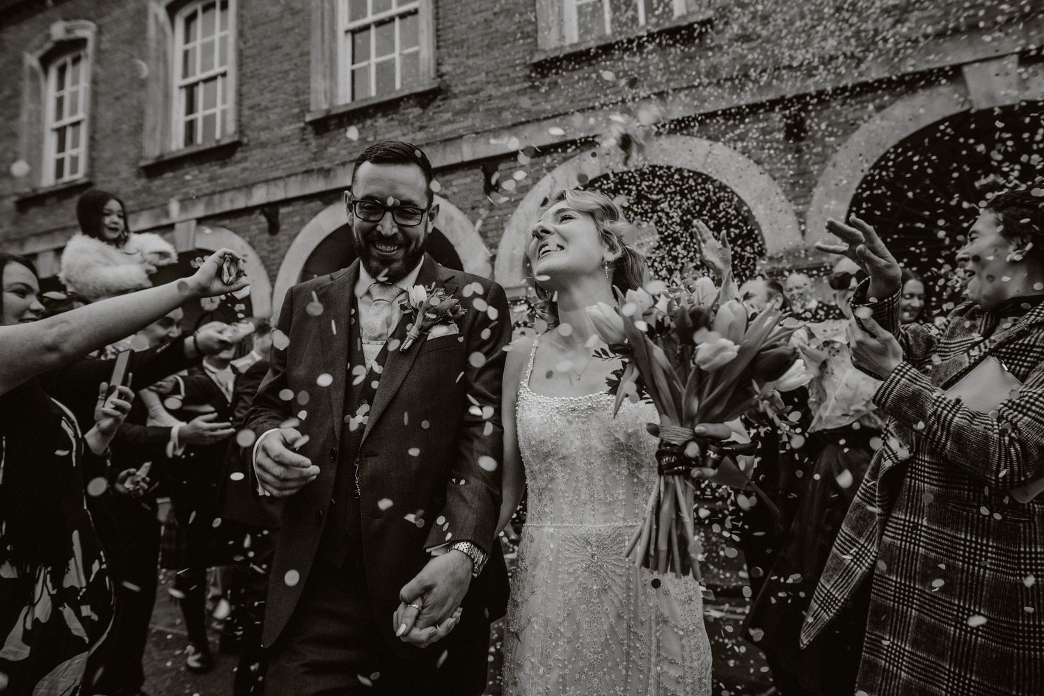 A joyful wedding couple walks through a shower of confetti outside a historic brick building with arched windows. The George Rye