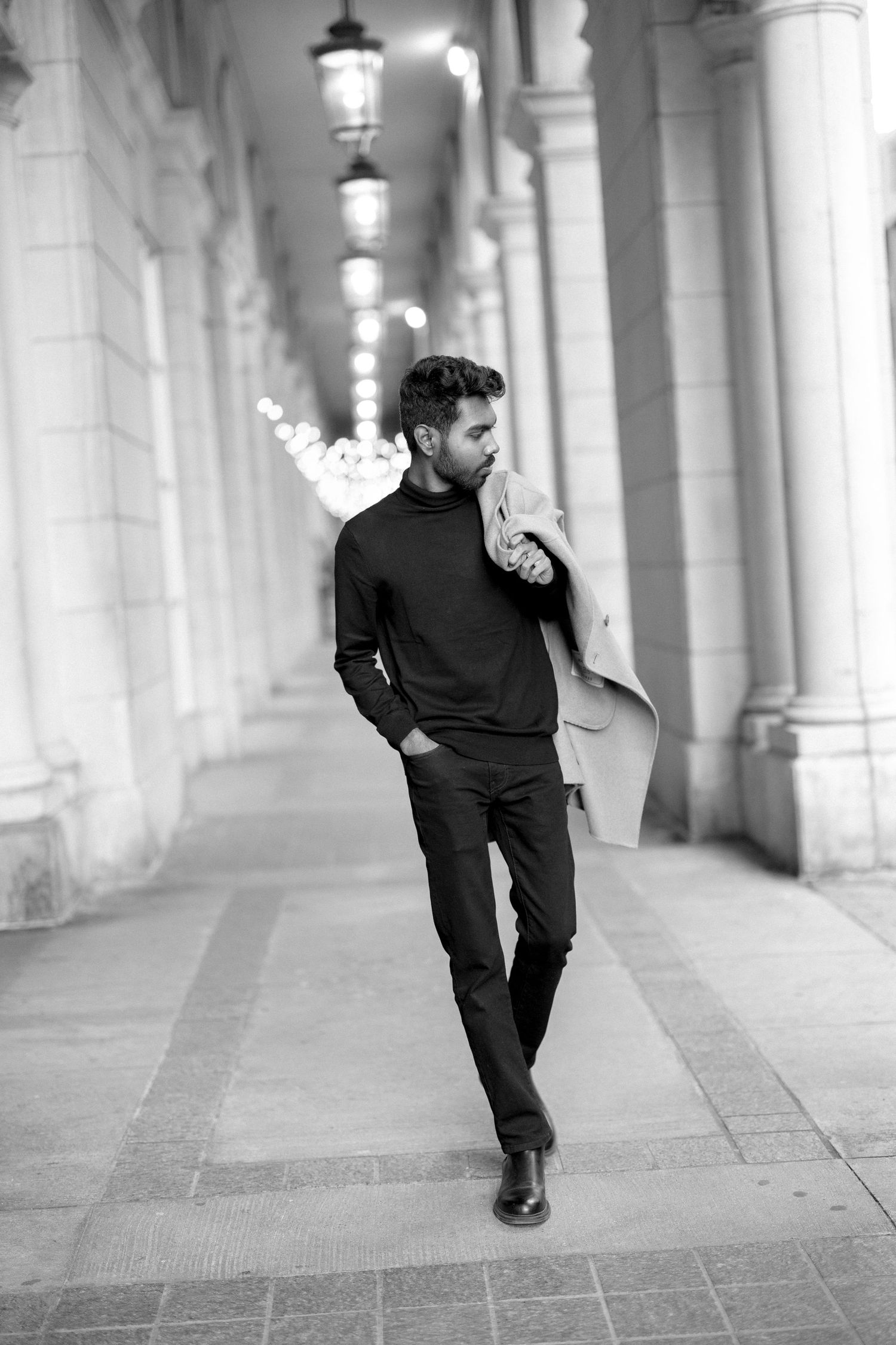 Stylish black and white portrait of a person in dark attire walking along a colonnade.