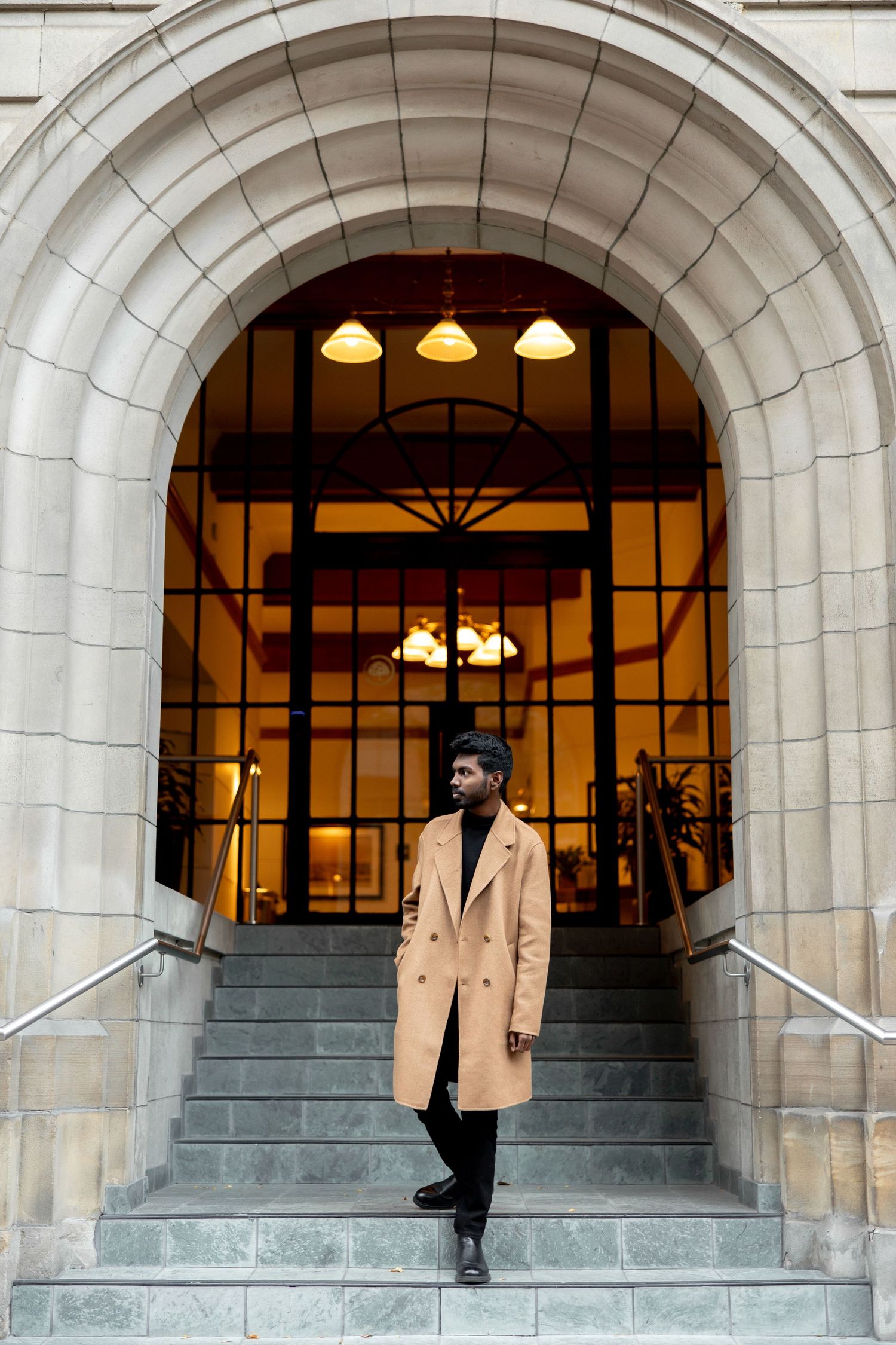 A person in a camel coat poses on stone steps at an elegant archway entrance.