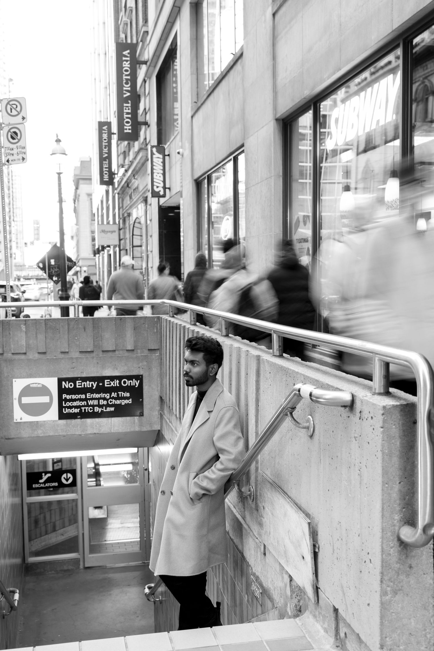 Black and white street scene with a person in a light coat near subway entrance steps.
