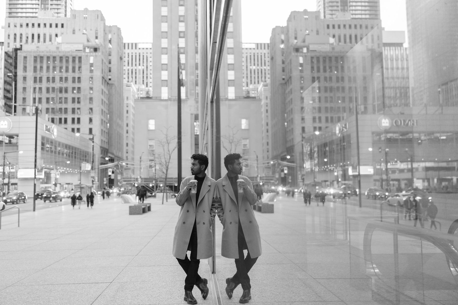 Black and white cityscape reflection showing people in winter coats amid tall urban buildings in New York City.