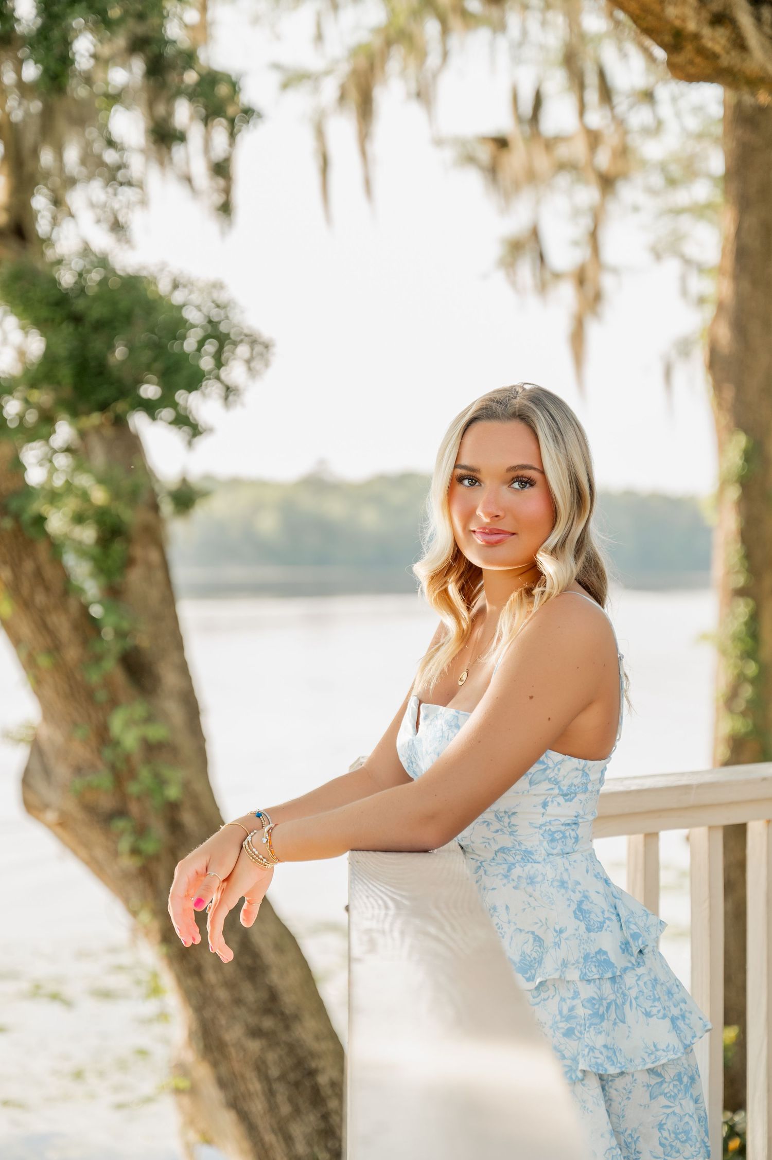 Figure in blue floral dress poses on wooden deck with Spanish moss trees and water in background.
