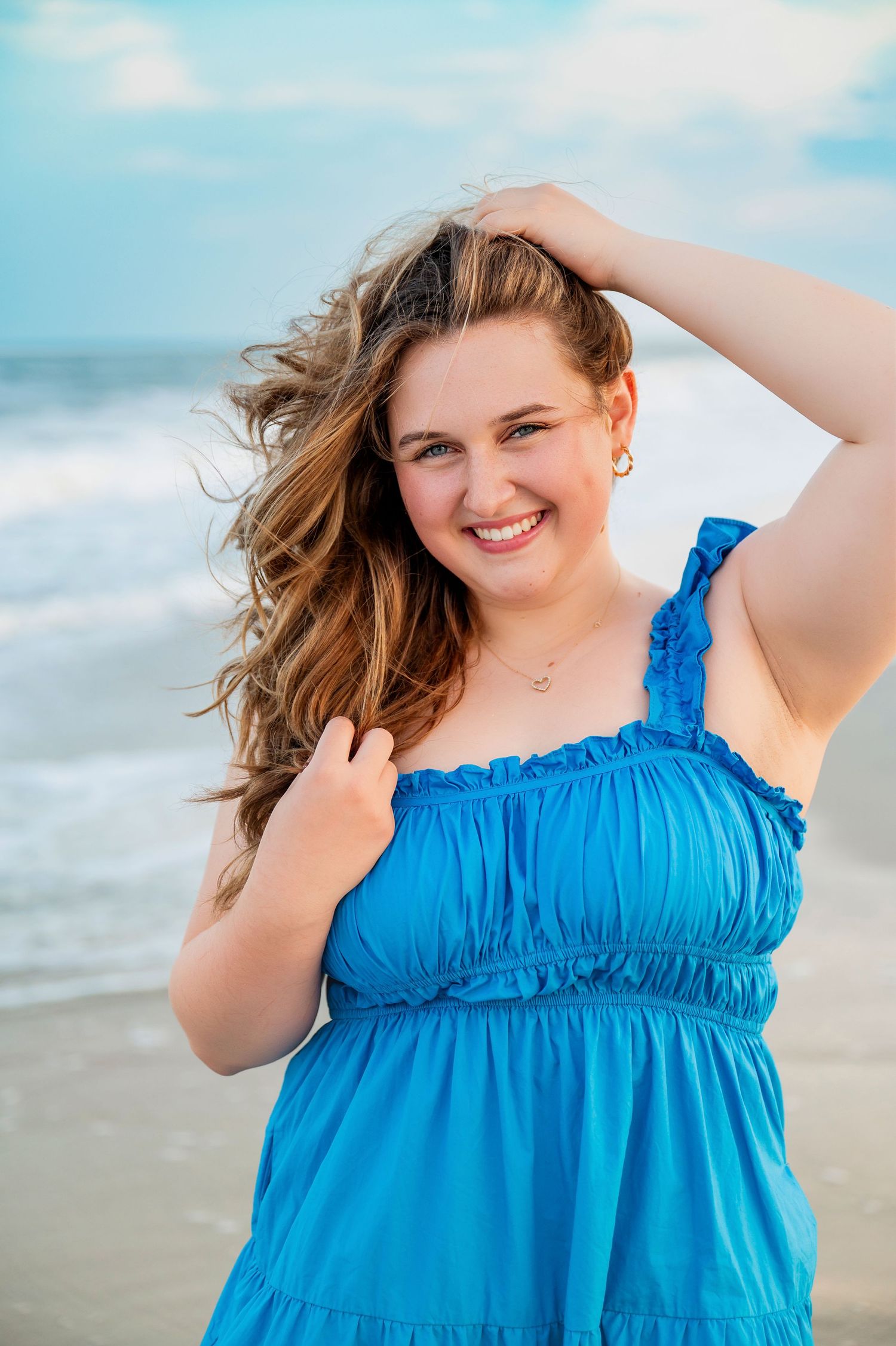 Person in bright blue summer dress poses joyfully on beach with wind-swept hair.