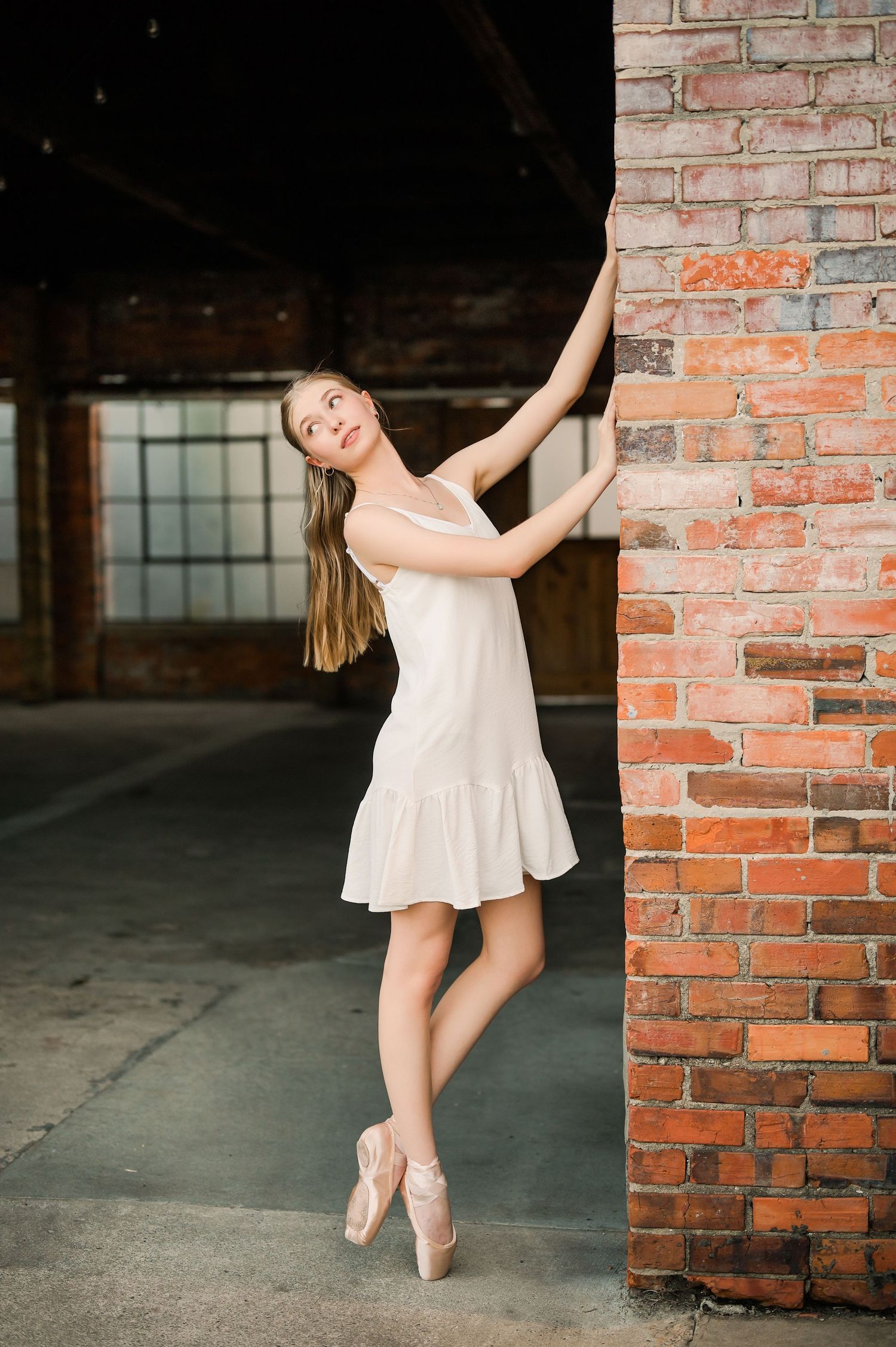 Dancer in white dress and ballet pointe shoes poses gracefully against brick wall.
