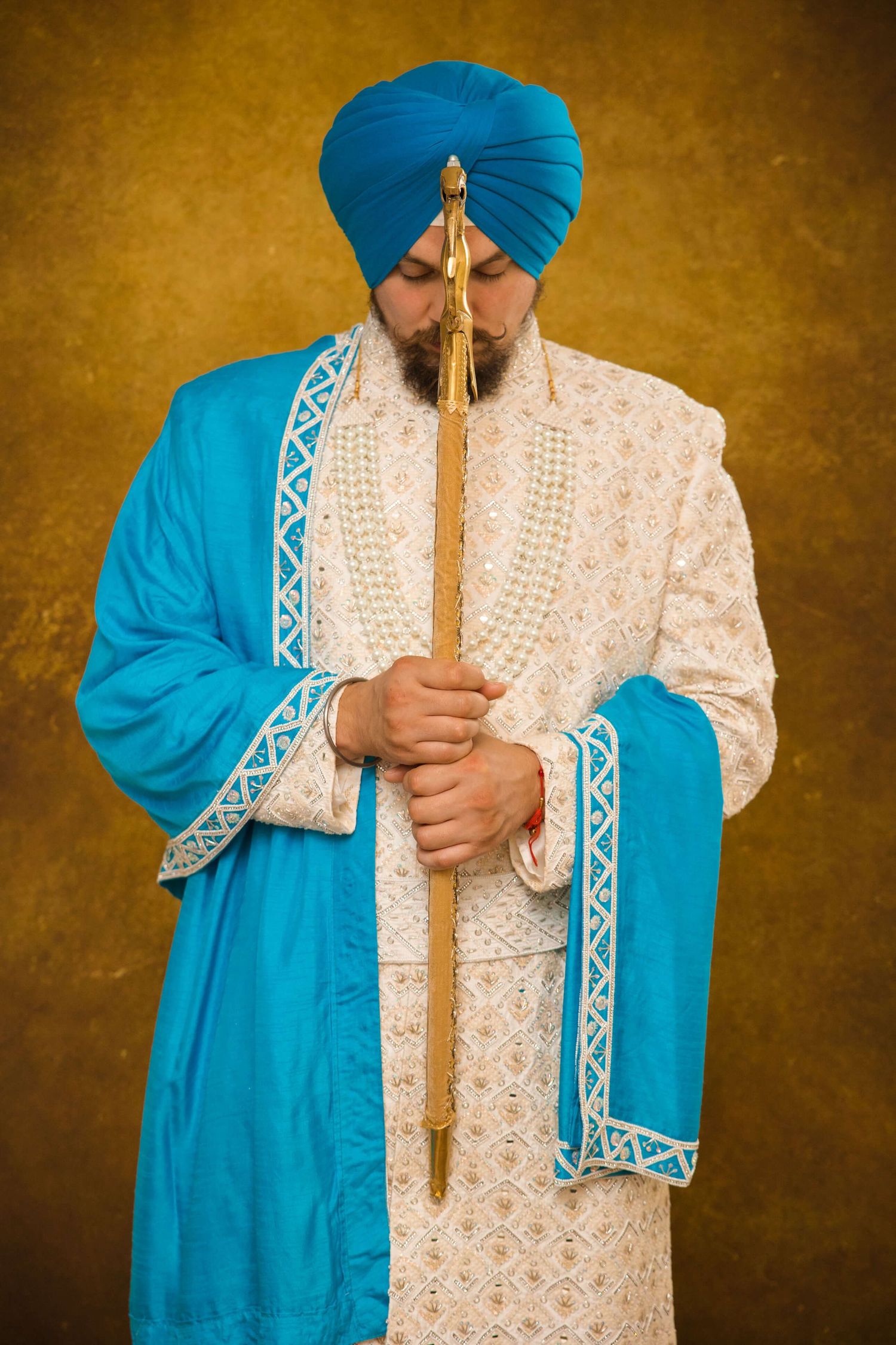 A Sikh groom posing in front of a kirpan, wearing a white luxury Kurta