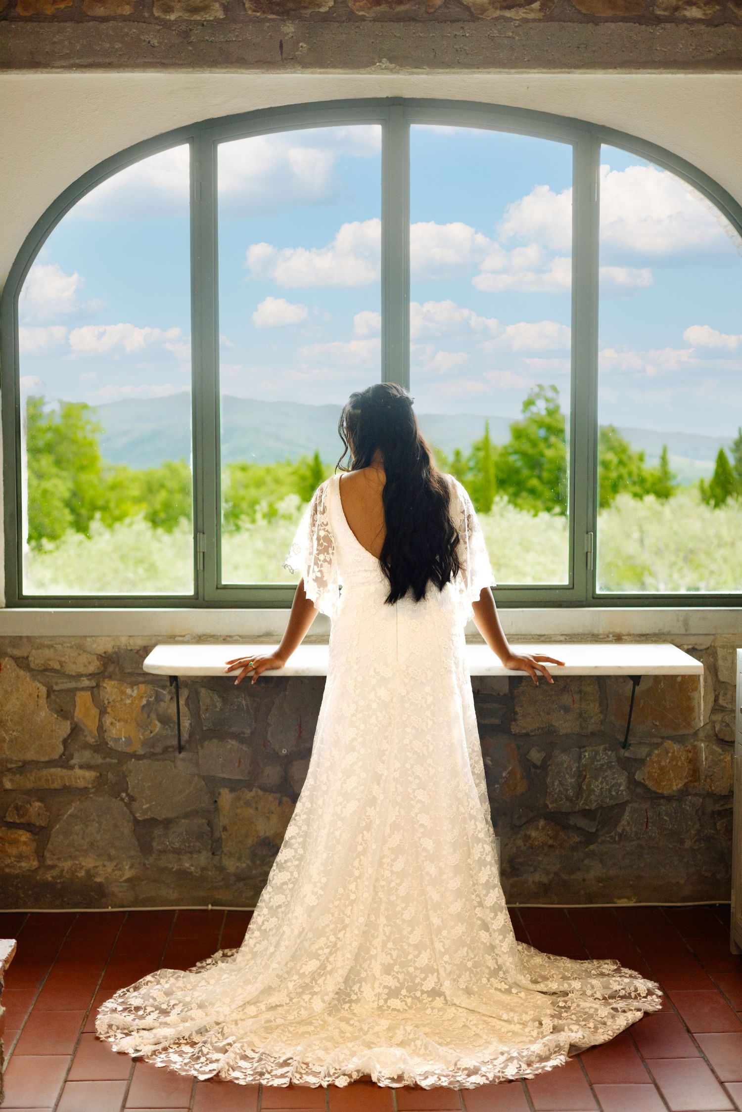 Bride in lace wedding gown gazes out arched window overlooking scenic mountain view.
