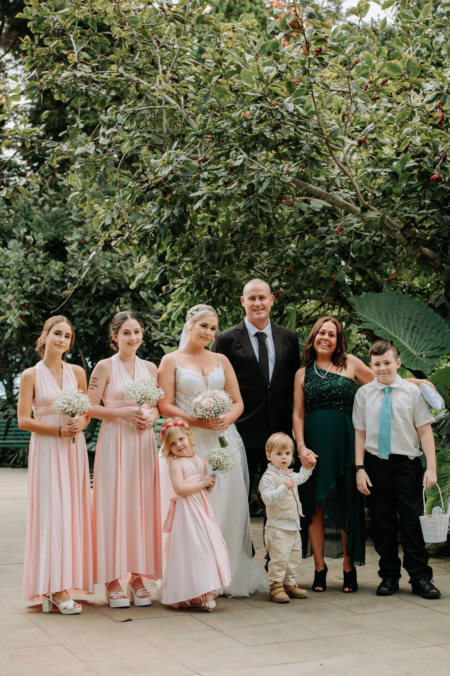 Wedding party in pink and formal attire posing together under a leafy tree.