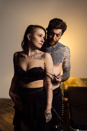A couple in dark clothing with visible tattoos pose intimately against a warm-lit backdrop.
