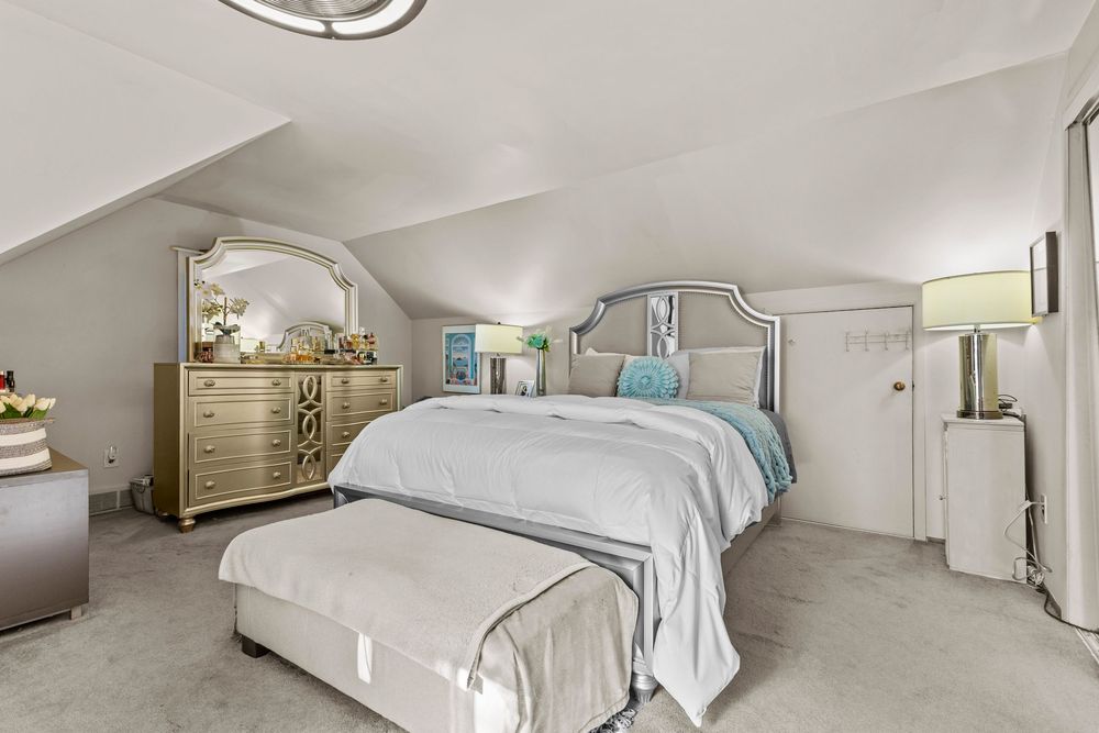 Cozy attic bedroom with sloped ceilings, plush carpet, elegant bedding, and gold dresser with mirror.