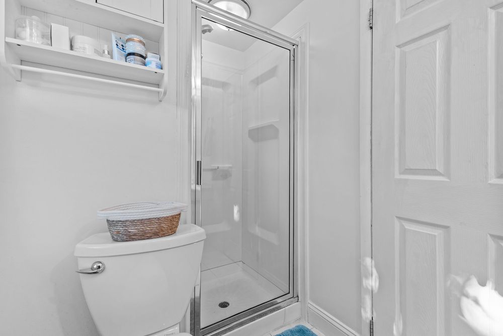 White bathroom with glass shower stall, toilet, and wall-mounted shelving unit.