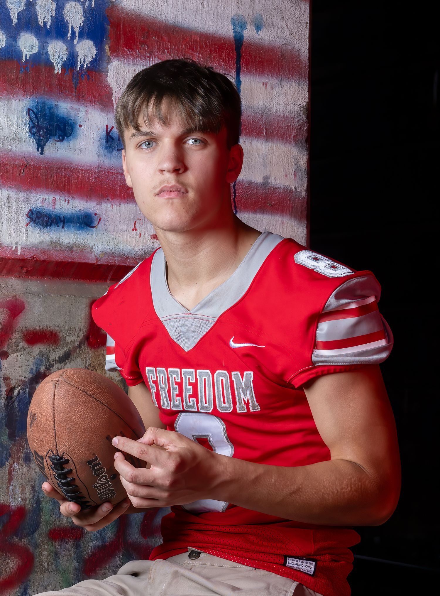 A high school athlete in a red Freedom jersey poses with football in front of American flag graffiti.