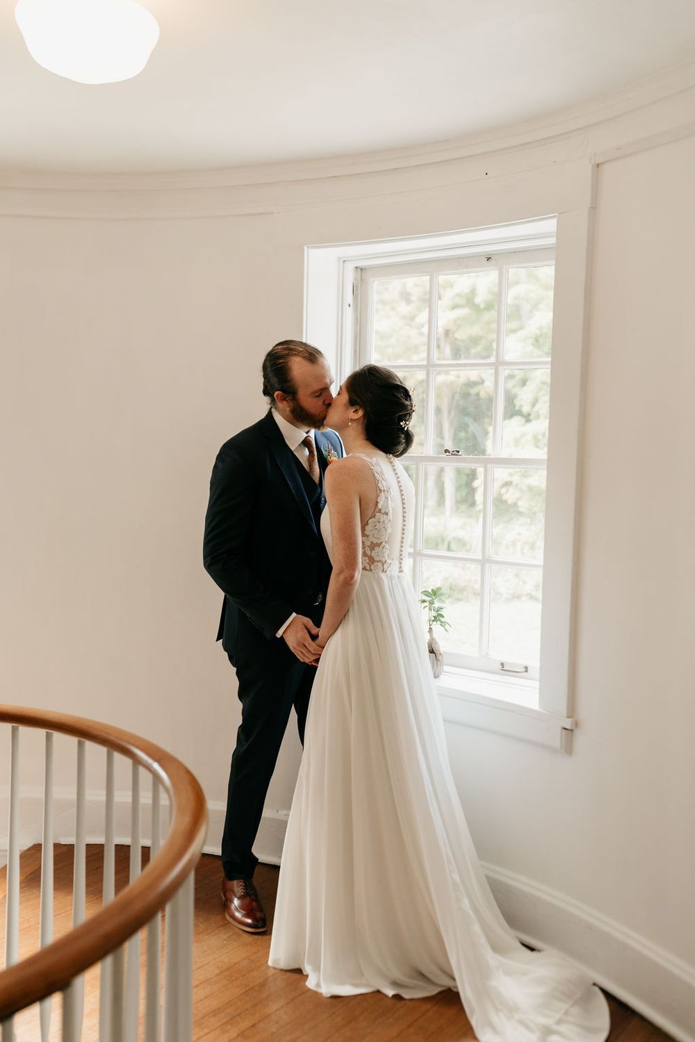 A tender wedding moment between a couple in formal attire standing by a bright window in a white interior space.