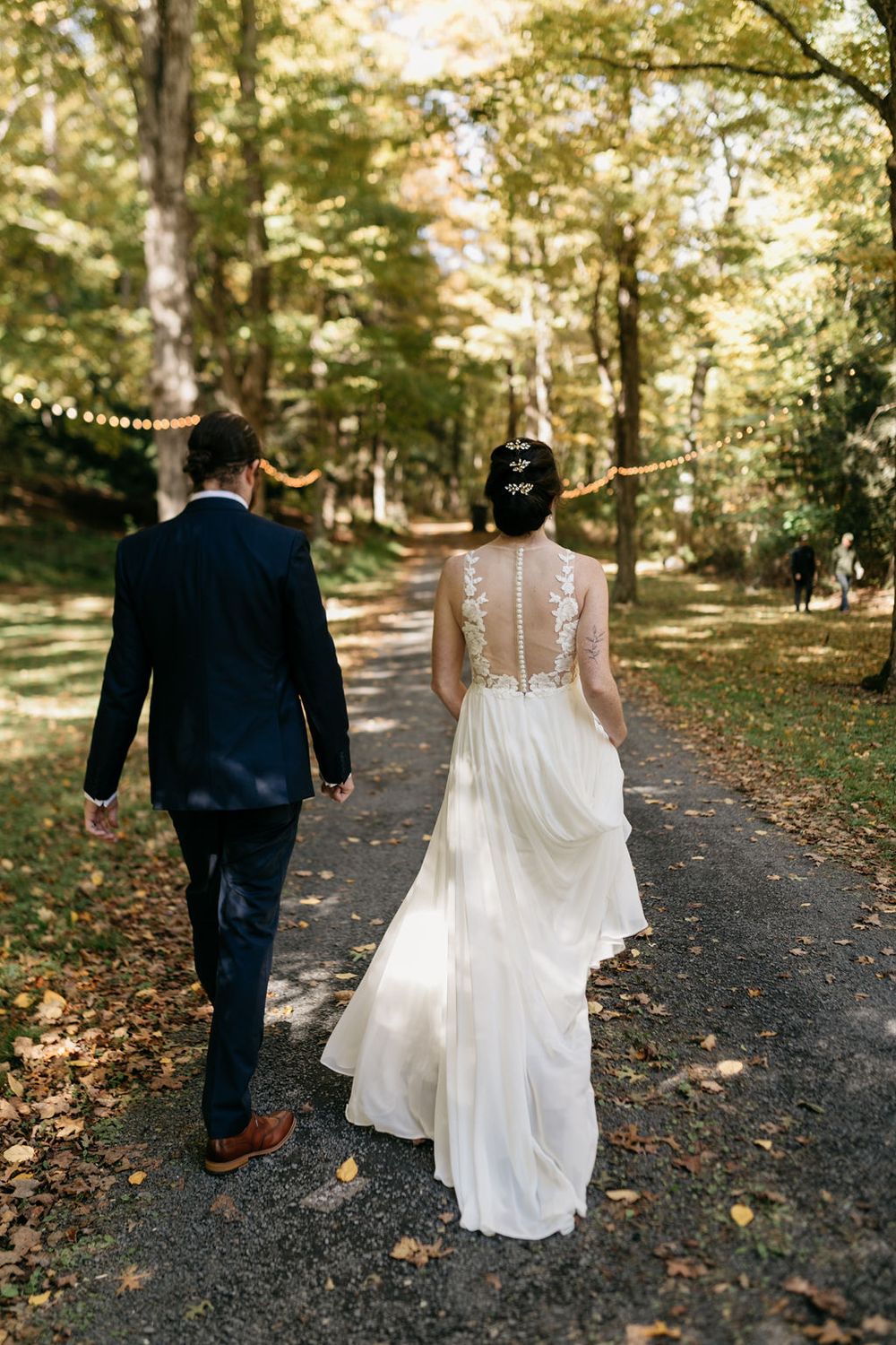 A bride and groom walk down a tree-lined path with autumn leaves scattered on the ground in golden sunlight.