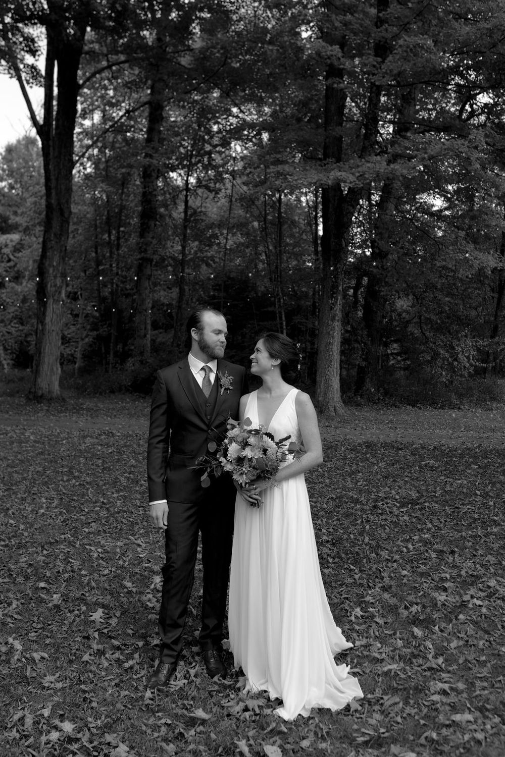 A romantic wedding couple poses together on fallen autumn leaves in a wooded setting.