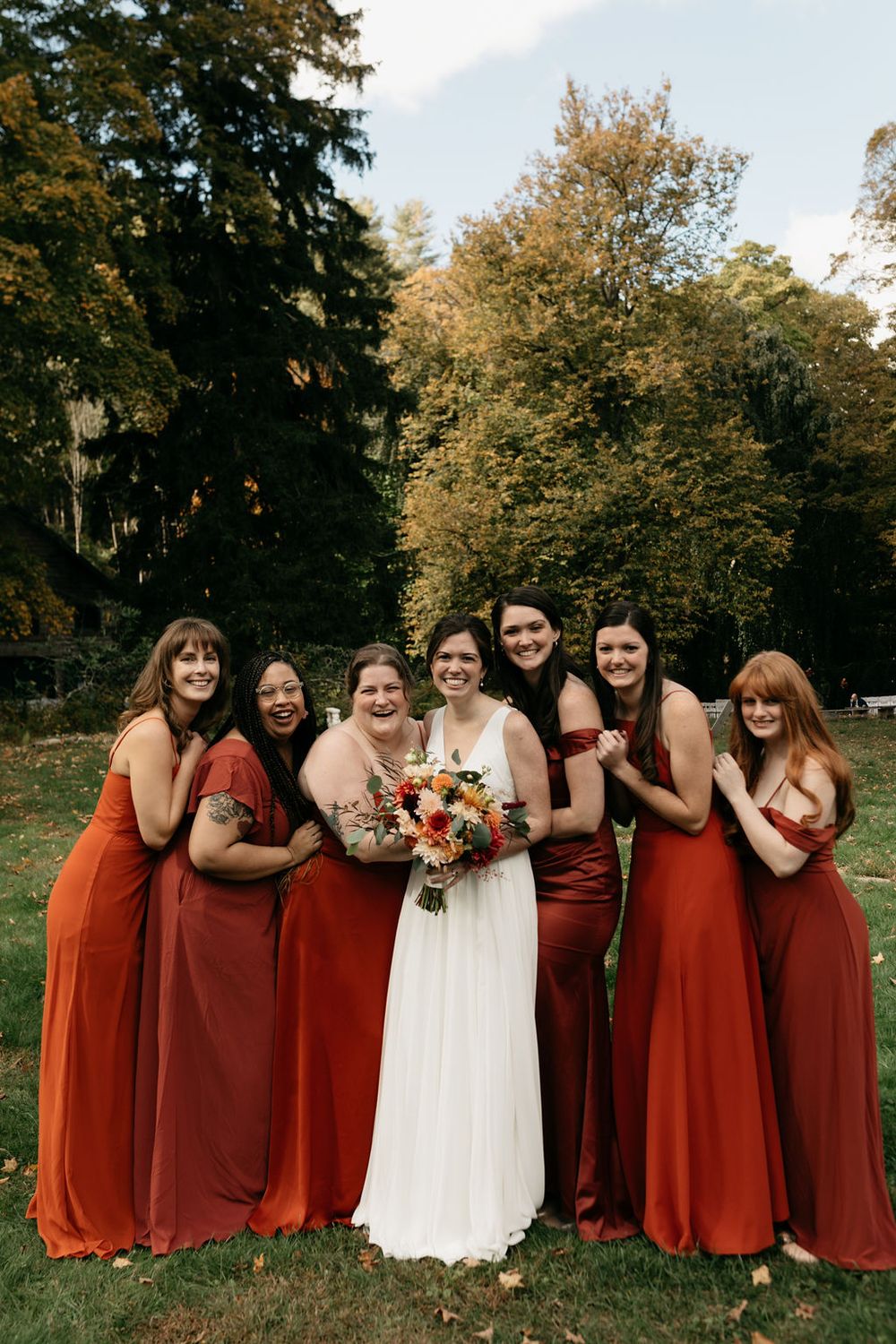 A wedding party in burgundy bridesmaid dresses poses together on a lawn with trees in the background.