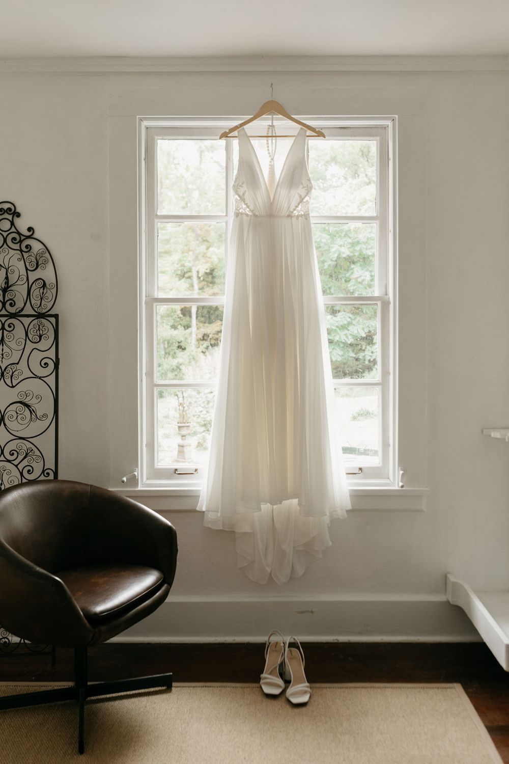 An elegant white wedding dress hangs in front of a bright window with white heels placed beneath.