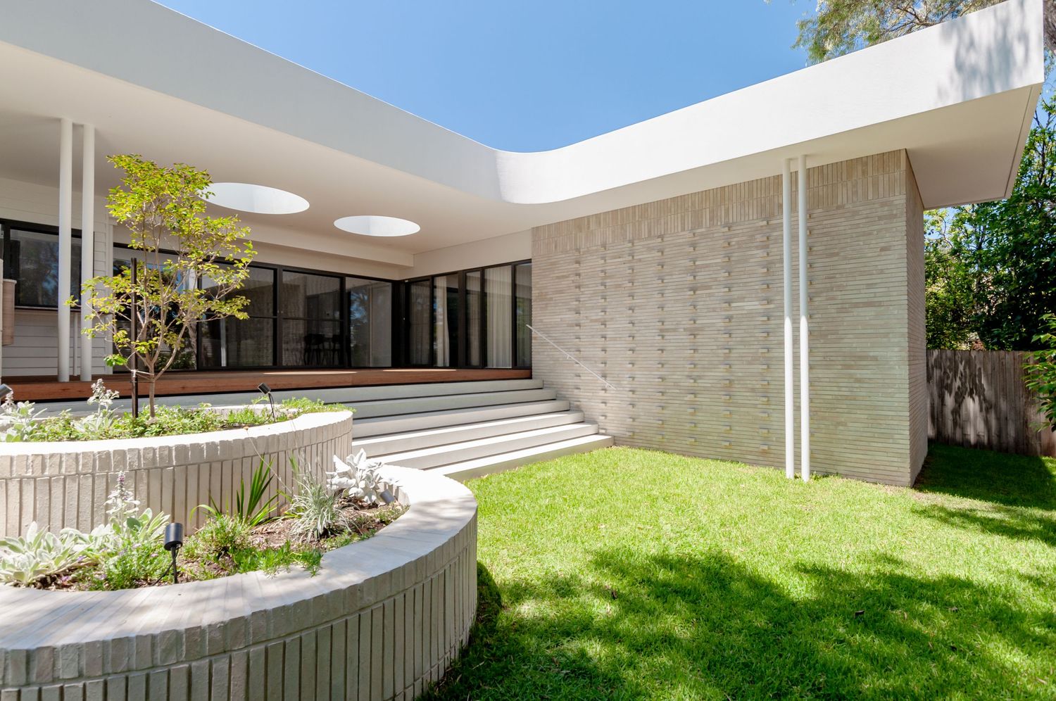 Modern white mid-century house exterior with curved stone planters and manicured lawn in bright sunlight.
