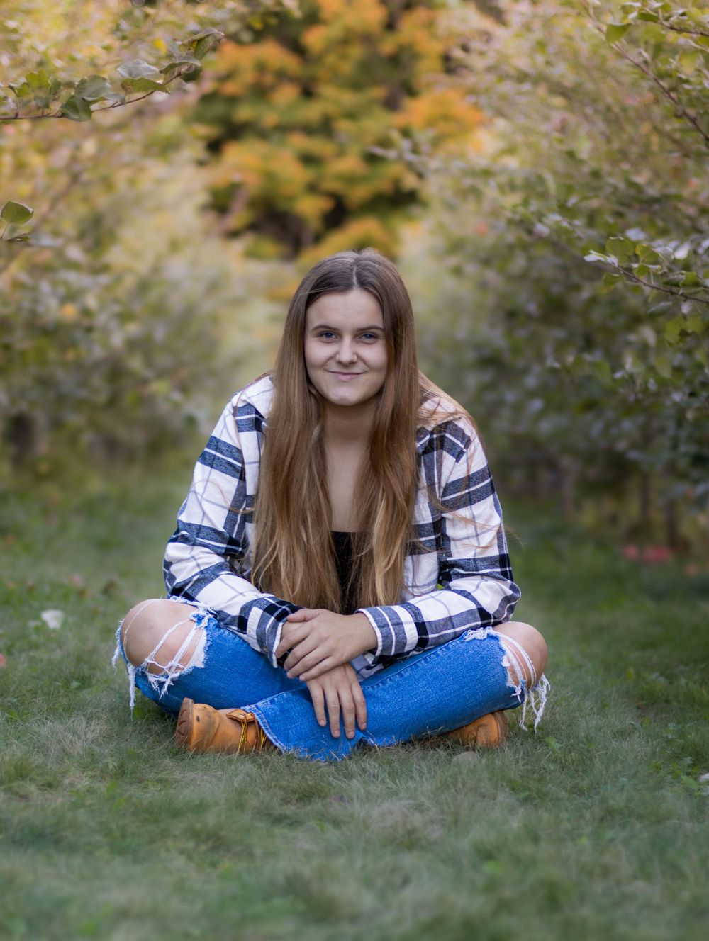 Person in plaid shirt and jeans sits casually on grass during autumn photoshoot.