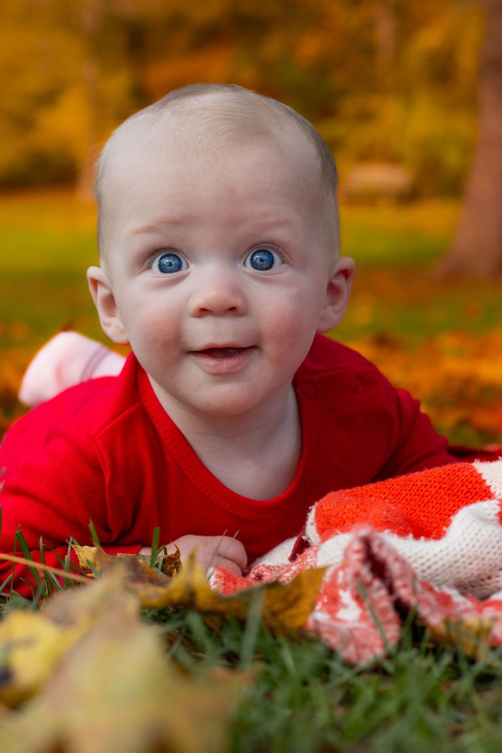 A baby wearing red lies on fallen autumn leaves with a bright expression.