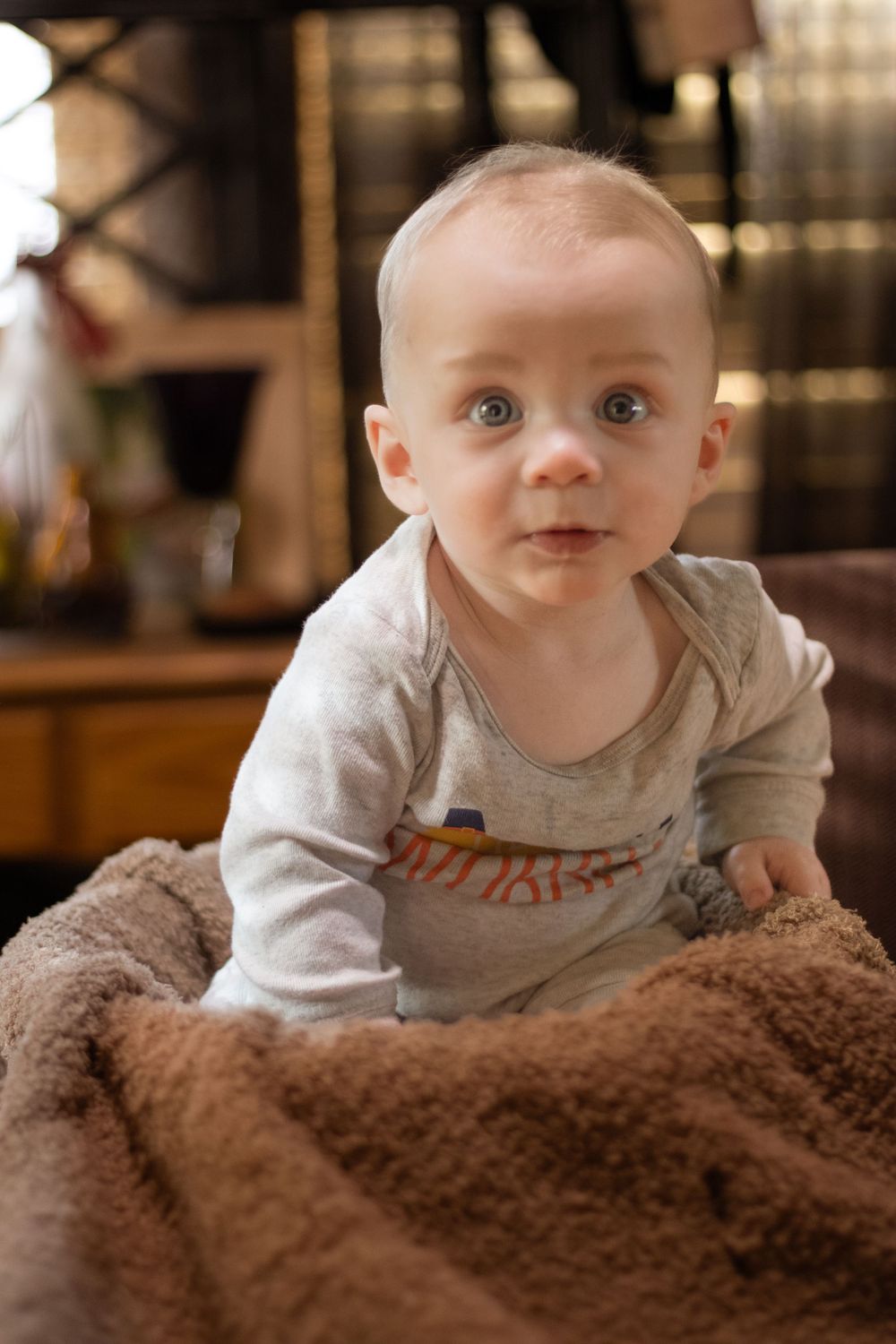 A baby in a gray shirt lies on a soft brown blanket while looking up with bright eyes.