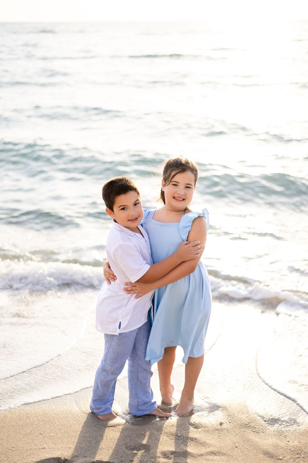 Two figures in coordinating blue and white outfits share a warm embrace on a sunlit beach.