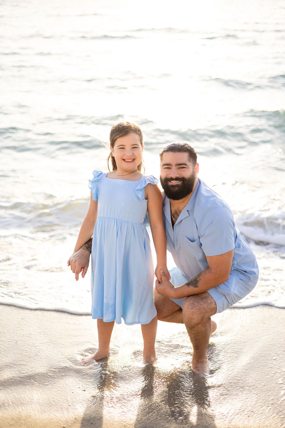 A heartwarming beach portrait shows two people in blue outfits standing at the ocean's edge during sunset.