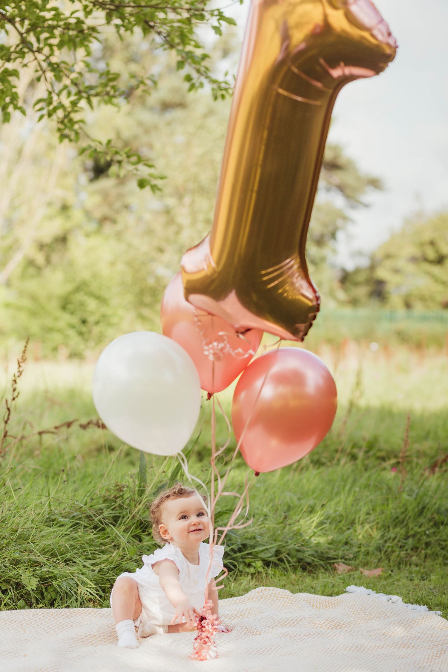 A baby celebrating their first birthday with gold and pink balloons on a blanket outdoors.