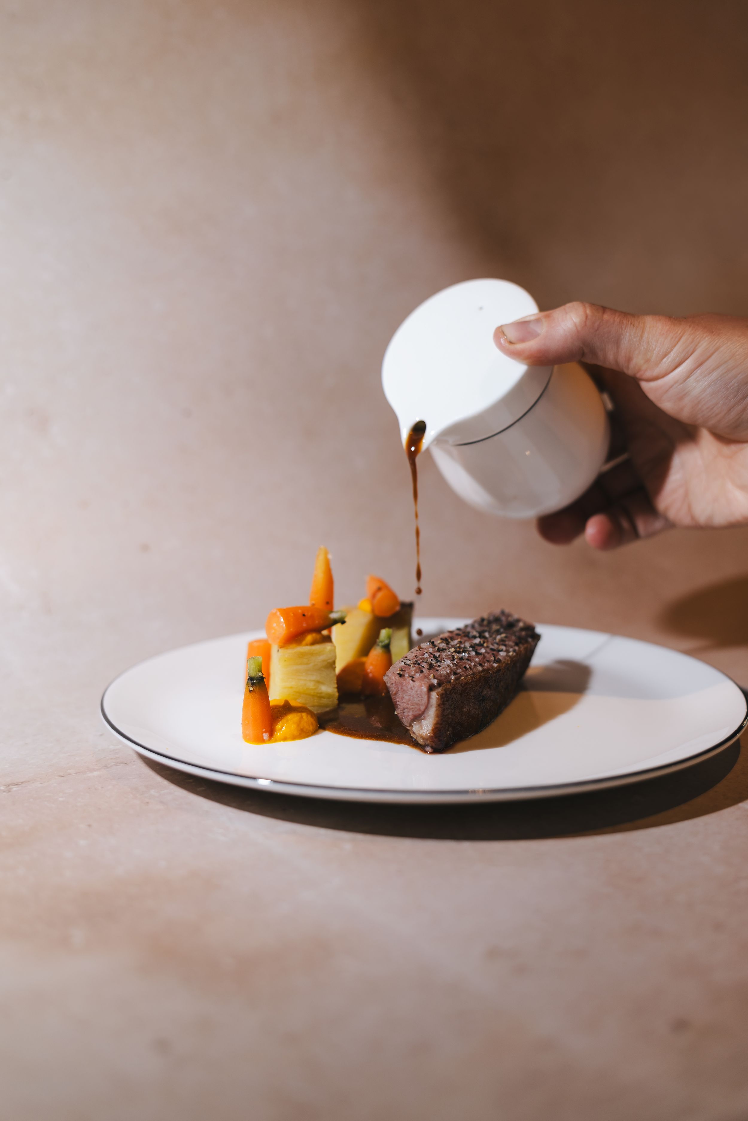 Hand pouring rich brown sauce over a gourmet beef dish with roasted carrots and vegetables on a white plate.