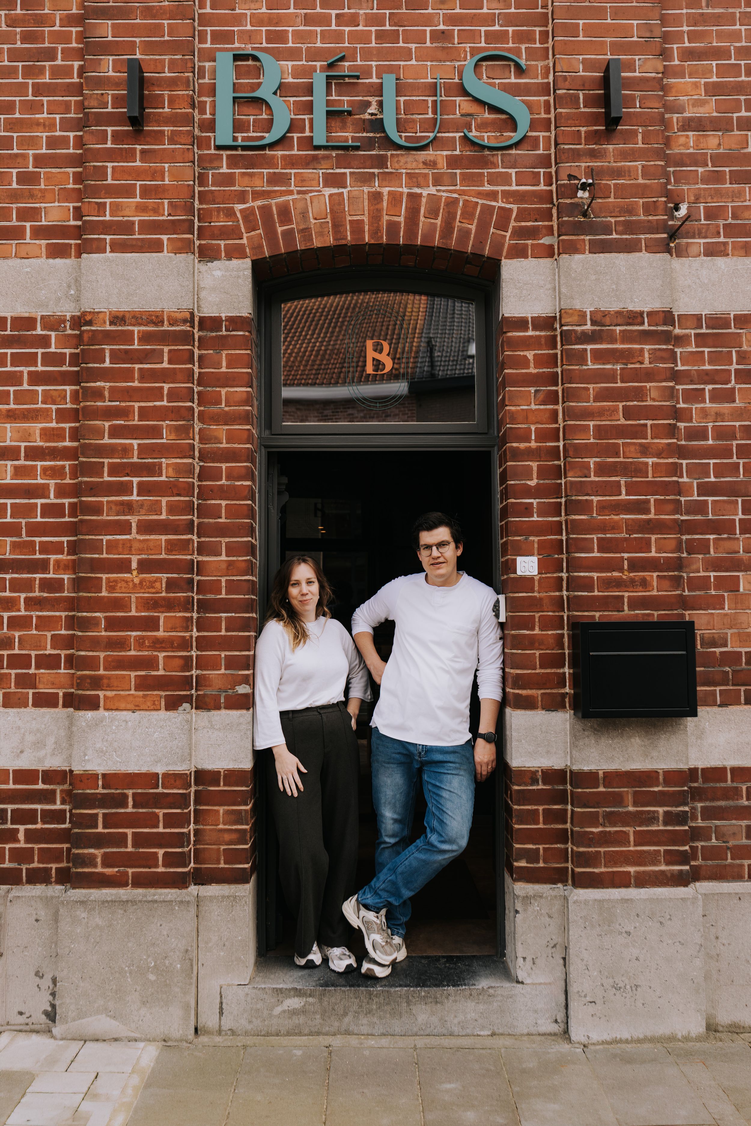 Two people smiling in the doorway of Béus restaurant, a charming red brick building with vintage signage.